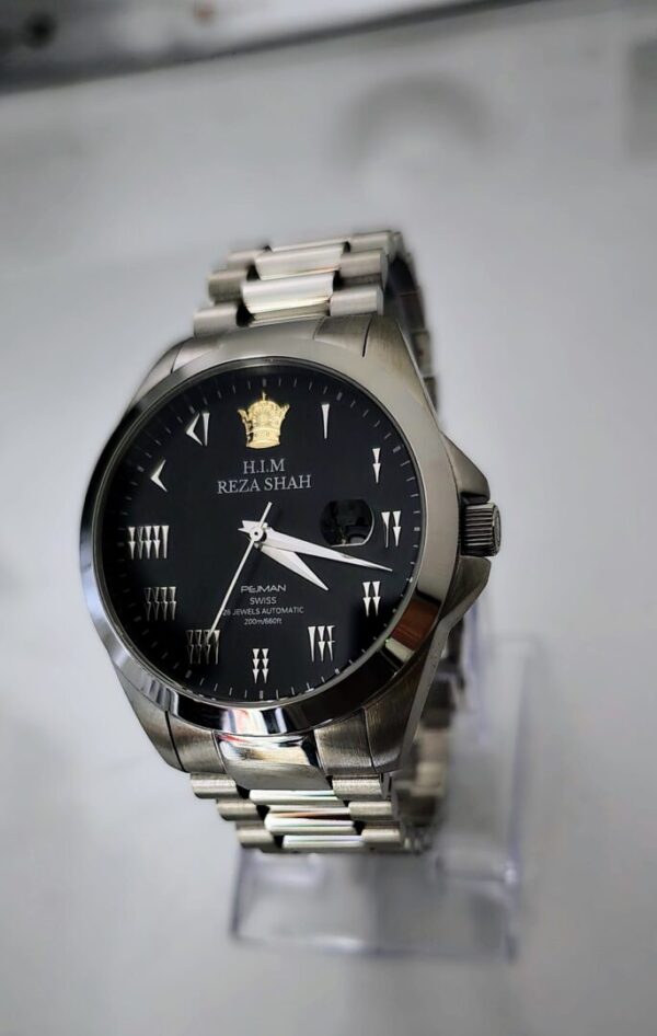 A black watch is on display in front of a white wall.