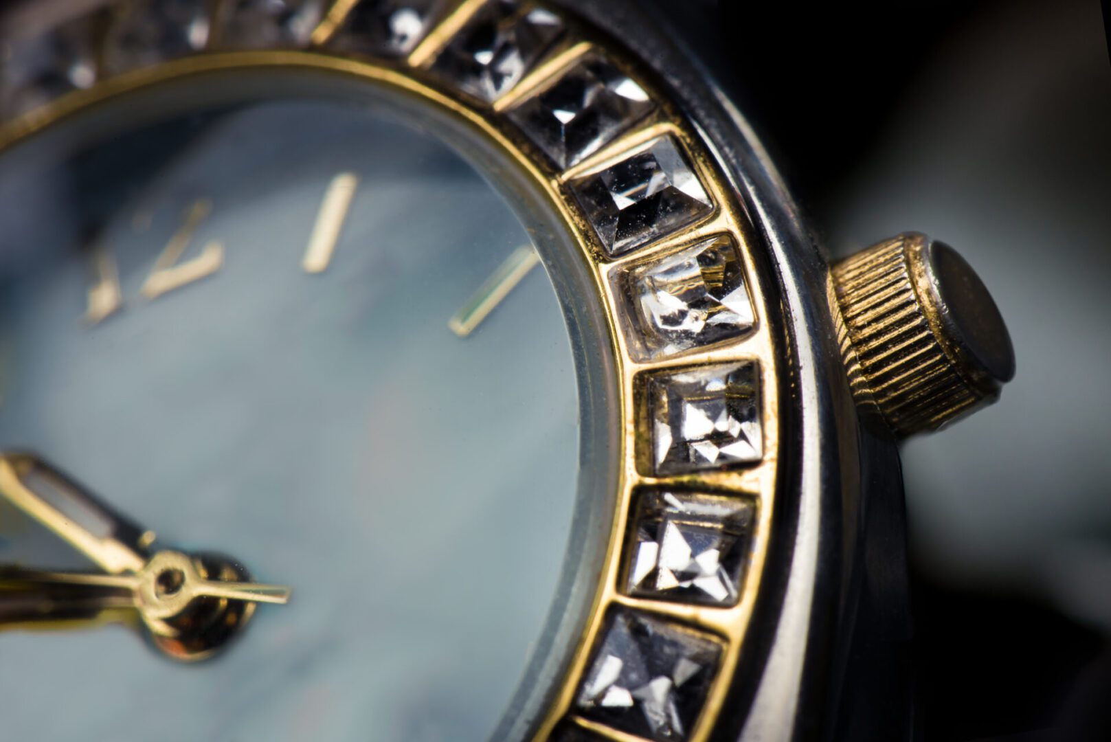 Close-up of a watch face with gold accents and a crystal-studded bezel. The watch hands point to 12 and 2, and the crown is visible on the right.